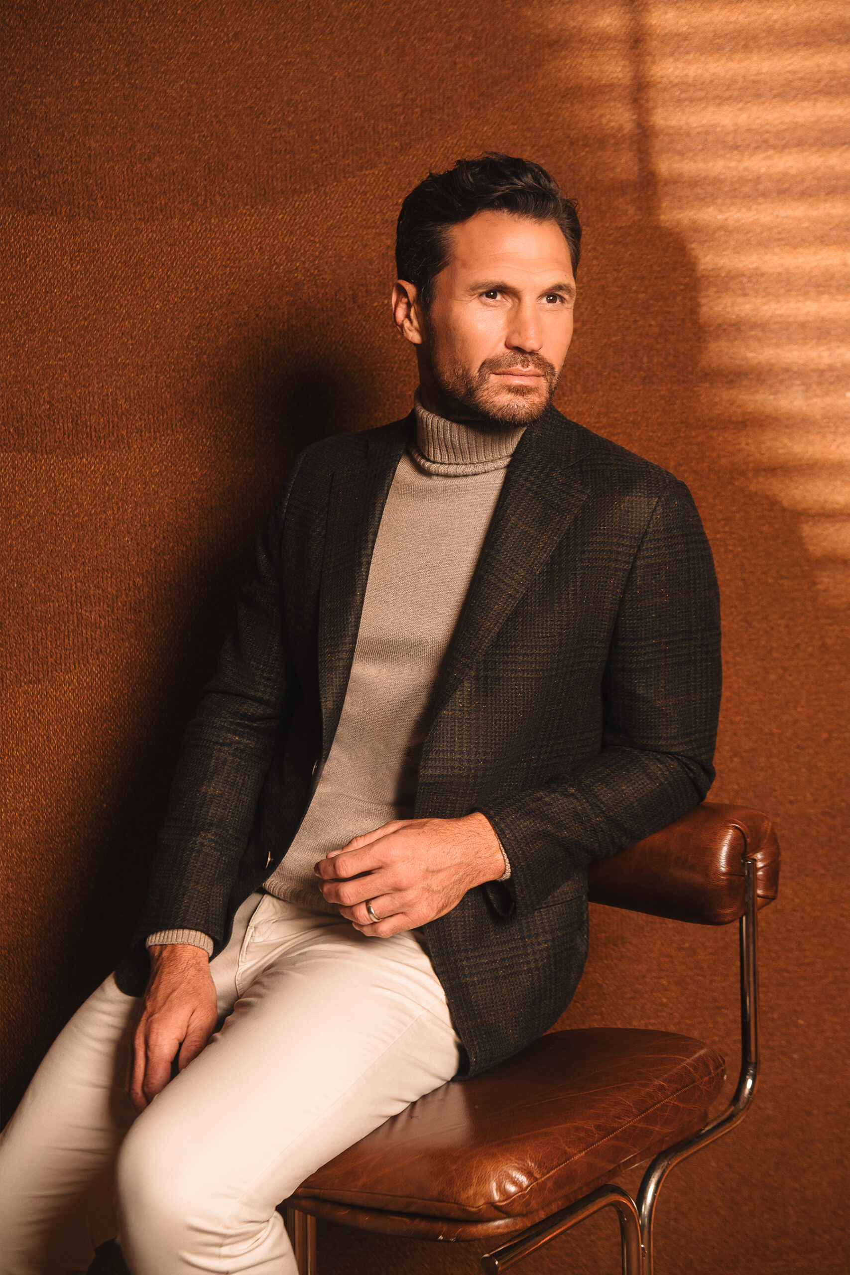 Model wearing a dark checked jacket and taupe turtleneck against a warm brown background